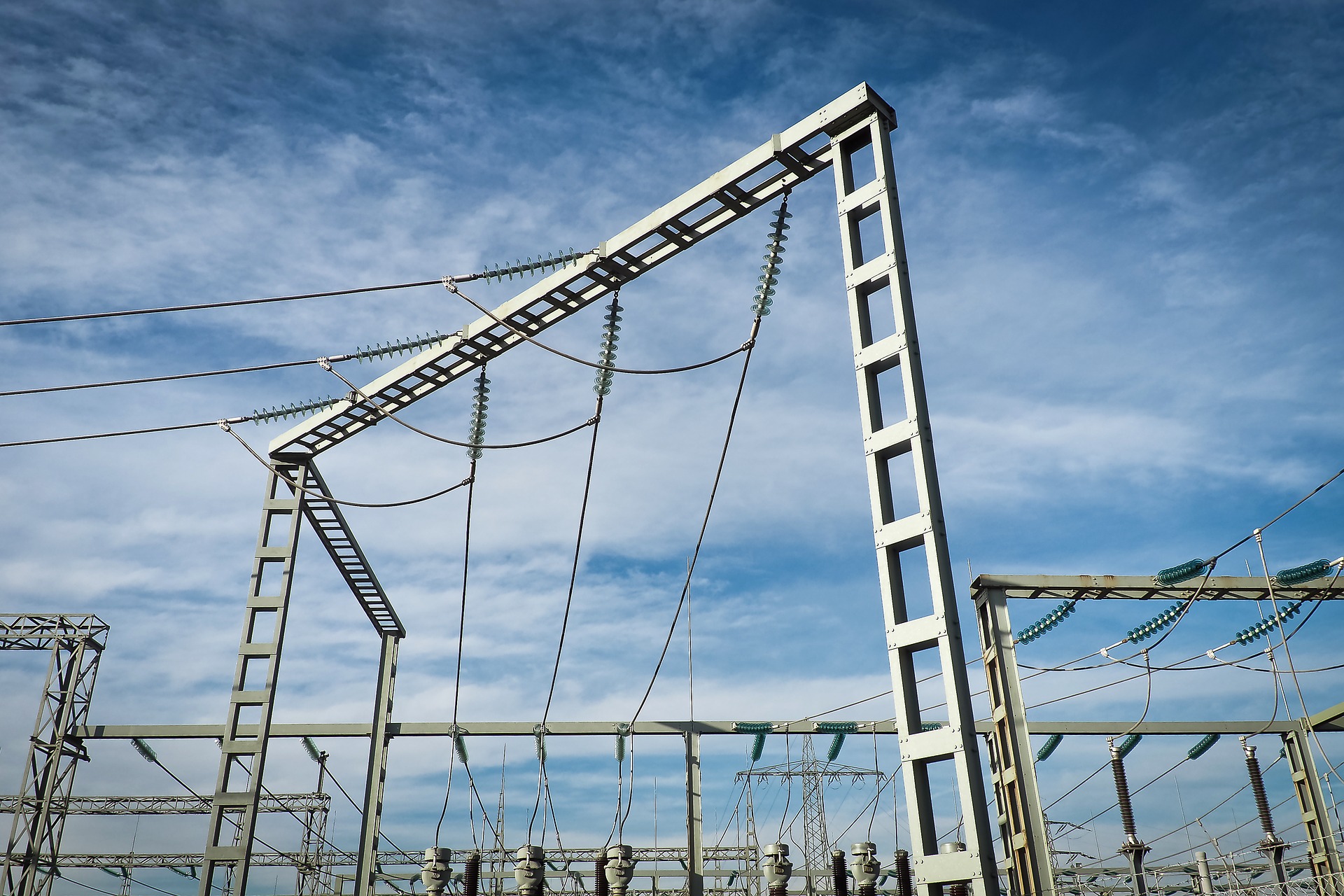A metal structure from an electrical substation is shown against a blue sky. The image features power lines and insulators that are part of the electricity distribution system, emphasizing the infrastructure essential for power transmission.