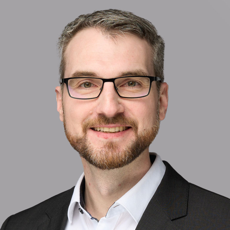 Portrait eines Mannes mit Brille und gepflegtem Bart, der lächelt. Er trägt ein helles Hemd und einen dunklen Anzug. Der Hintergrund ist grau.