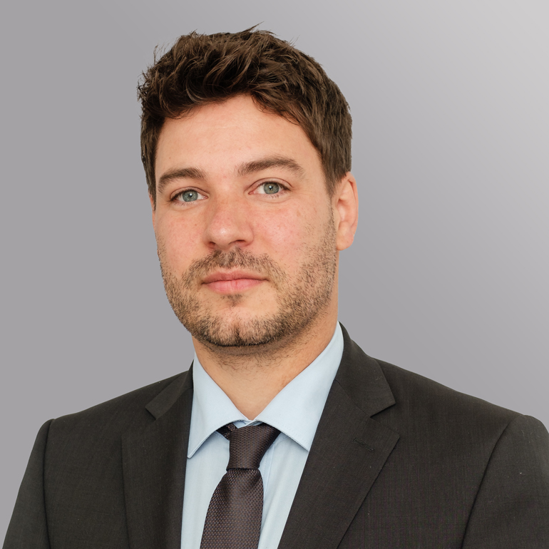 Porträt eines Mannes mit kurzen, dunklen Haaren und Bart. Er trägt einen schwarzen Anzug, ein hellblaues Hemd und eine dunkelgraue Krawatte. Der Hintergrund ist in sanften Grautönen gehalten. Der Mann blickt direkt in die Kamera mit einem neutralen Ausdruck.