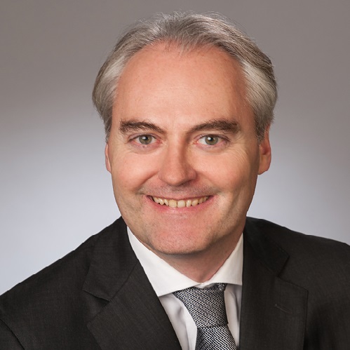 Portrait eines Mannes mit kurzen, grauen Haaren und einem freundlichen Lächeln. Er trägt einen dunklen Anzug und eine hellere Krawatte. Der Hintergrund ist neutral und verstärkt den Fokus auf sein Gesicht.