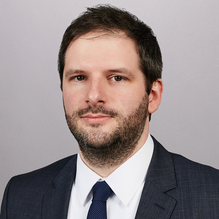 Ein Mann mit dunkelbraunem Haar und Bart trägt einen dunklen Anzug und eine weiße Hemdbluse mit einer blauen Krawatte. Er schaut direkt in die Kamera vor einem grauen Hintergrund.