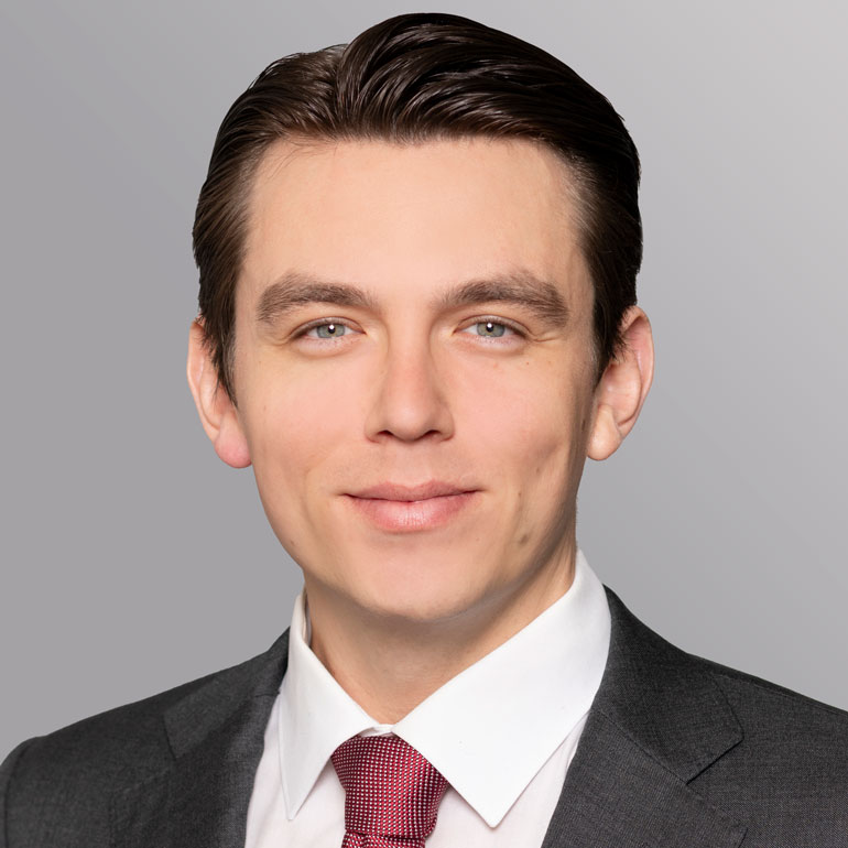 Porträt eines jungen Mannes mit kurzem, glattem Haar. Er trägt einen dunklen Anzug und ein weißes Hemd mit roter Krawatte. Der Hintergrund ist grau. Der Mann lächelt freundlich und schaut direkt in die Kamera.