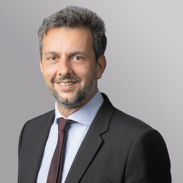 Ein lächelnder Mann mit grauem Haar trägt einen schwarzen Anzug und ein hellblaues Hemd mit einer dunklen Krawatte. Der Hintergrund ist einhellig grau.