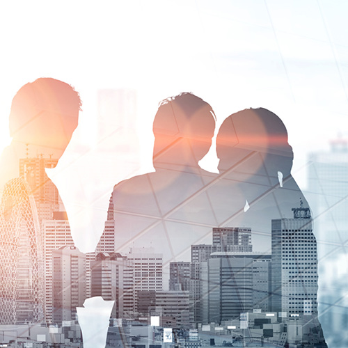 Eine Gruppe von drei Personen steht zusammen, umgeben von einer Silhouette einer Stadtlandschaft. Die Konturen der Menschen sind verschwommen, und das Bild vermittelt einen Eindruck von Zusammenarbeit und urbaner Umgebung. Das Licht hinter ihnen schafft eine sanfte Atmosphäre.