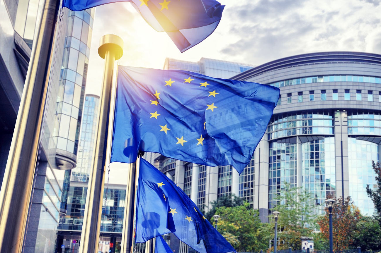 The image shows several European Union flags waving on poles in front of a modern building. The flags are blue with a circle of yellow stars, symbolizing unity among member states. The scene is illuminated by sunlight, creating a vibrant atmosphere.