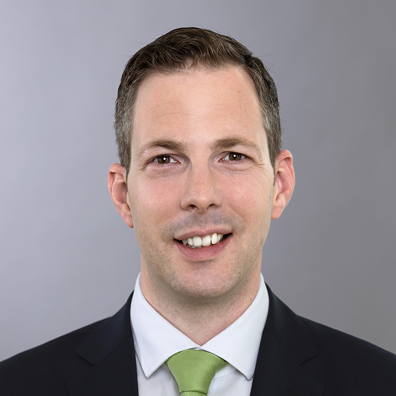 Ein Mann mit kurzen, dunklen Haaren lächelt in die Kamera. Er trägt einen dunklen Anzug mit einer hellblauen Hemd und einer grünen Krawatte. Der Hintergrund ist grau und neutral gehalten.