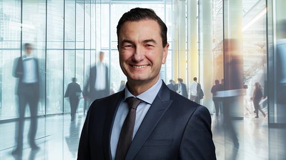 Ein lächelnder Mann im Anzug steht im Vordergrund. Im Hintergrund sind unscharfe Silhouetten von Menschen in einem modernen, hellen Gebäude zu sehen, was eine geschäftliche Umgebung vermittelt.