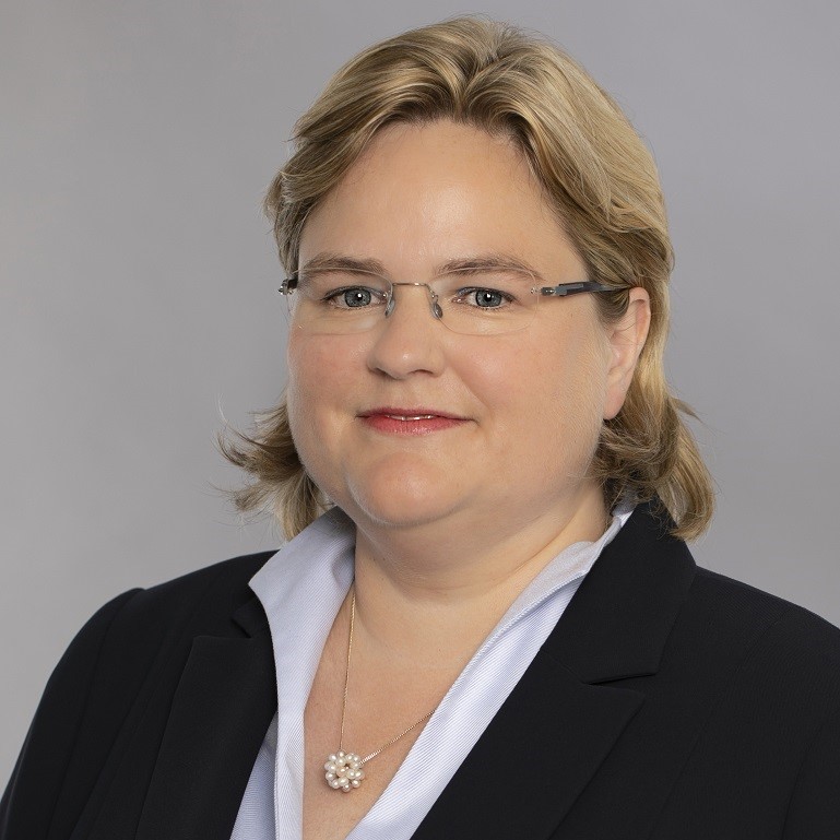 Eine blonde Frau mit schulterlangen Haaren trägt eine Bluse und einen Blazer. Sie schaut direkt in die Kamera und hat eine freundliche Miene. Im Hintergrund ist eine neutrale, hellgraue Wand sichtbar.