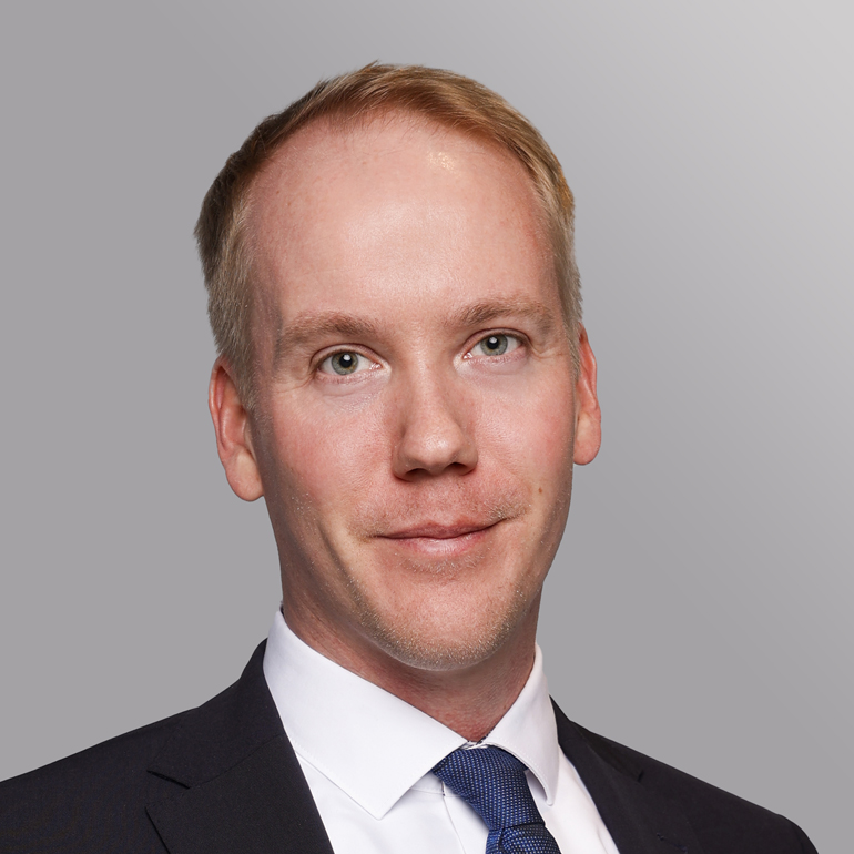 Ein Mann mit blonden Haaren und einem leichten Bart lächelt in die Kamera. Er trägt einen dunklen Anzug und eine weiße Hemd mit einer blauen Krawatte. Der Hintergrund ist grau.