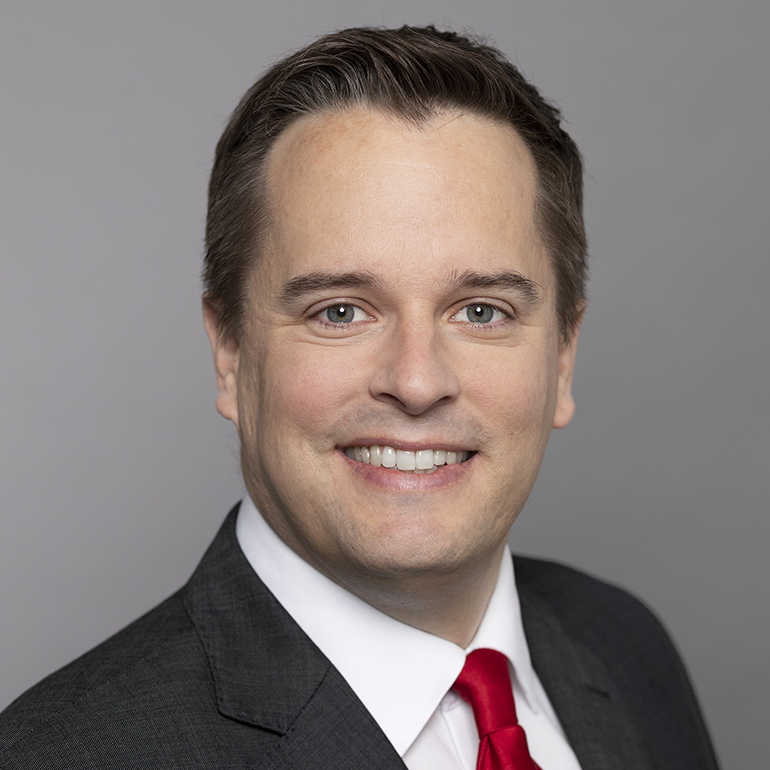 Porträt eines lächelnden Mannes mit kurzen Haaren, der einen dunklen Anzug und ein rotes Musterhemd trägt. Der Hintergrund ist einheitlich grau.