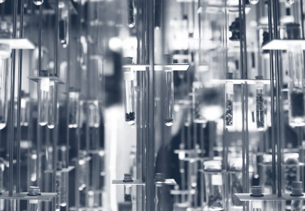 Ein transparenter Rahmen zeigt zahlreiche Glas-Röhrchen, die an Stangen hängen. Die Röhrchen sind teilweise mit kleinen Objekten gefüllt und reflektieren Licht. Die Anordnung schafft eine interessante visuelle Tiefe und stellt eine moderne wissenschaftliche Installation dar.