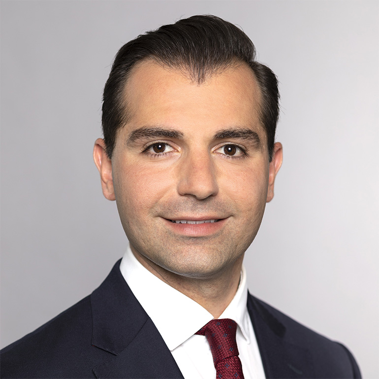 Ein junger Mann mit dunklem Haar und einem gepflegten Haarschnitt lächelt in die Kamera. Er trägt einen dunklen Anzug und eine rote Krawatte. Der Hintergrund ist hell und neutral gehalten, was den Fokus auf das Gesicht des Mannes lenkt.