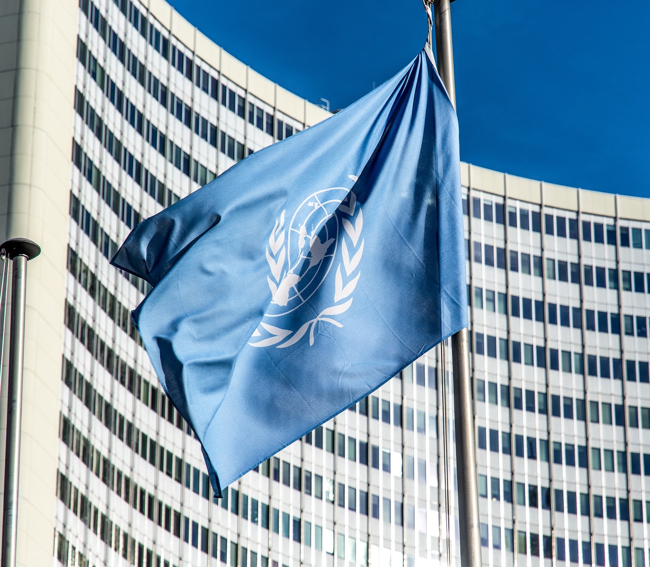 A blue flag featuring the emblem of the United Nations, which includes a world map surrounded by olive branches, is waving in the air. In the background, a modern building with a glass façade is partially visible under a clear blue sky.