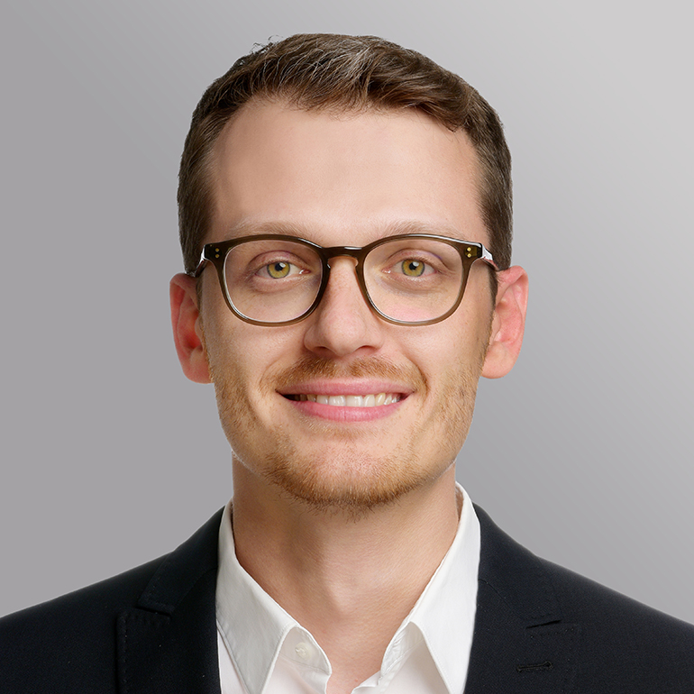 Ein junger Mann mit Brille lächelt freundlich in die Kamera. Er trägt ein schwarzes Jacket und ein weißes Hemd. Der Hintergrund ist grau und sorgt für einen professionellen Eindruck.