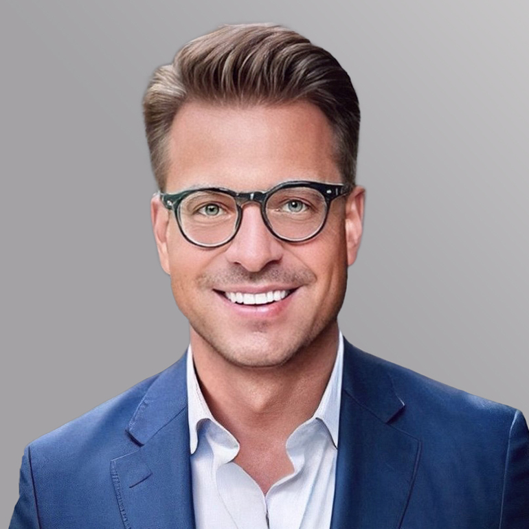 Ein lächelnder Mann mit kurzen, gestylten Haaren trägt eine Brille und einen blauen Anzug. Der Hintergrund ist grau. Sein freundlicher Gesichtsausdruck vermittelt Professionalität und Offenheit.