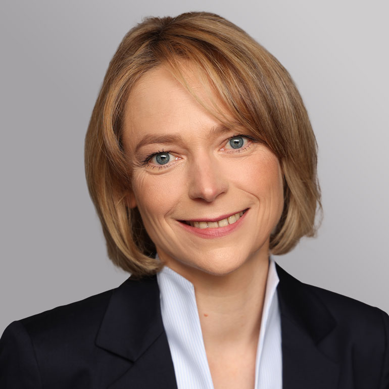 Porträt einer lächelnden Person mit kurzen, blonden Haaren. Sie trägt einen dunklen Blazer und ein helles Hemd. Der Hintergrund ist in sanften Grautönen gehalten. Die Person wirkt freundlich und professionell.