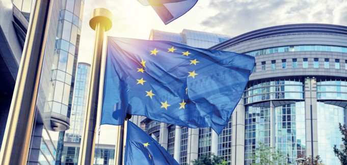 Die Flaggen der Europäischen Union wehen vor einem modernen Gebäude. Der Himmel ist teilweise bewölkt, und die Sonne scheint. Die blaue Flagge zeigt einen gelben Sternenumriss, ein Symbol für die EU.
