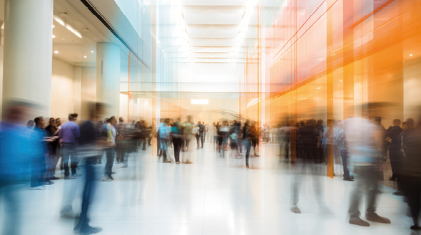 Das Bild zeigt eine belebte Lobby oder einen Foyer mit vielen Menschen, die sich bewegen. Die Personen erscheinen verschwommen, was auf eine lebhafte und dynamische Atmosphäre hinweist. Helle Innenarchitektur mit weichem Licht schafft eine einladende Umgebung.