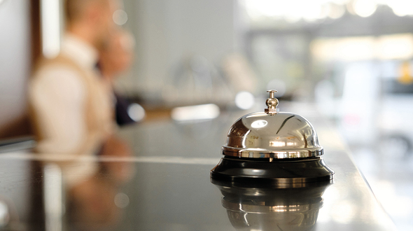 Ein glänzendes, silbernes Empfangsglockenläutchen steht auf einem dunklen Tisch. Im Hintergrund sind zwei verschwommene Personen zu sehen, wahrscheinlich Angestellte an einer Rezeption. Die Atmosphäre vermittelt einen professionellen Empfangsbereich.