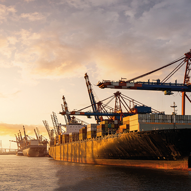 Containerfrachter liegen im Hafen, umgeben von Kränen, die Fracht laden und entladen. Die Szene wird von einem warmen Sonnenuntergang beleuchtet, der die Silhouetten der Schiffe und Kräne hervorhebt.