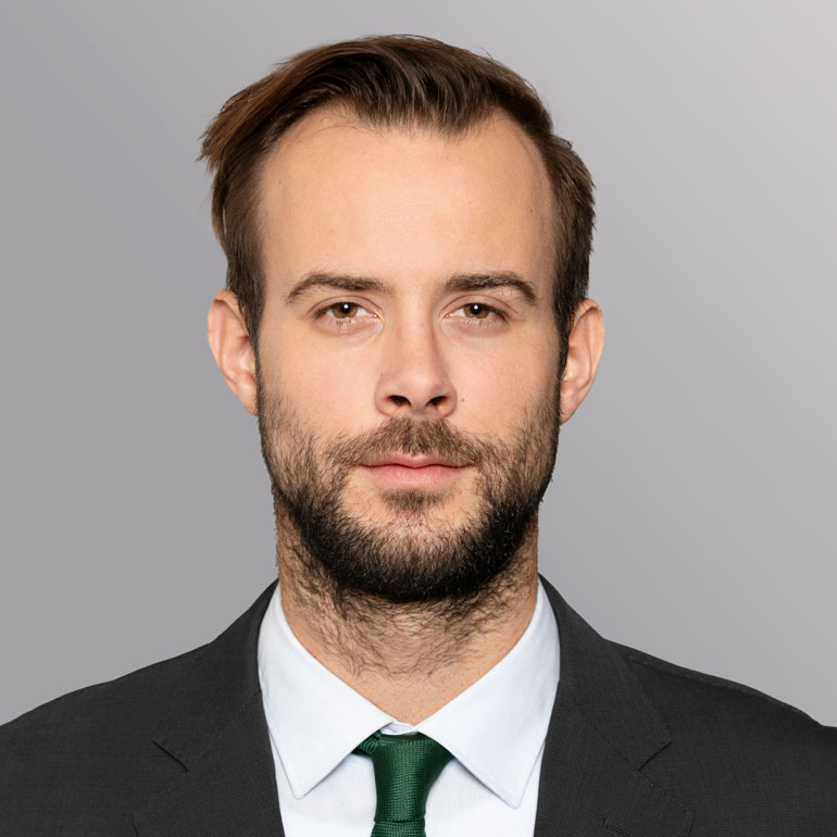 Ein junger Mann mit kurzem, braunem Haar und Vollbart schaut direkt in die Kamera. Er trägt einen dunkelgrauen Anzug mit einem weißen Hemd und einer grünen Krawatte. Der Hintergrund ist neutral grau.