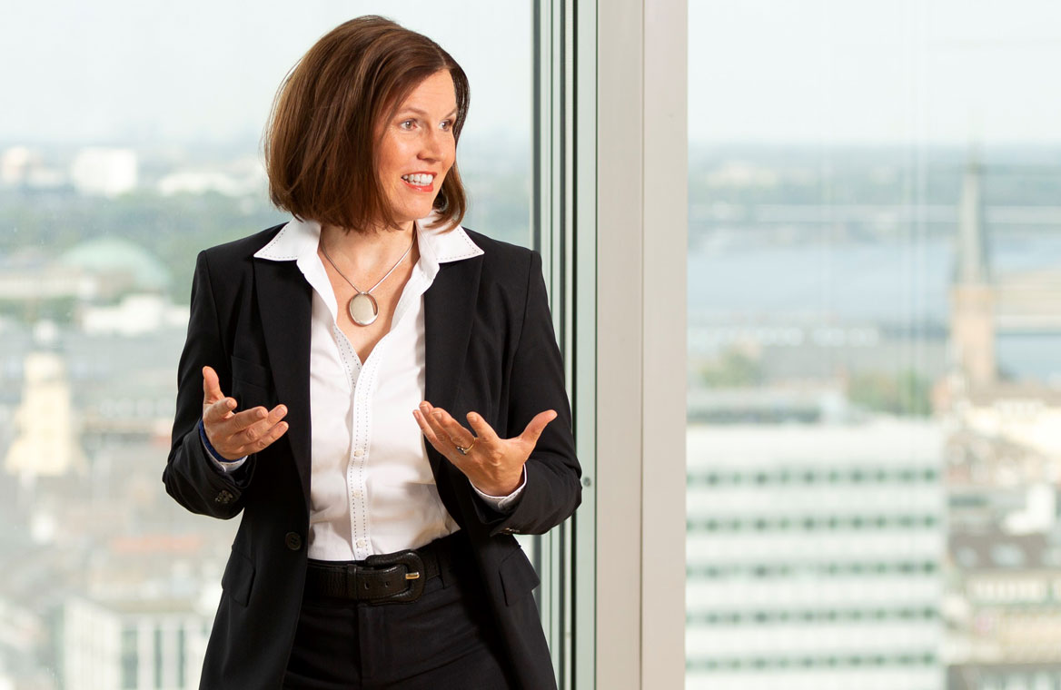 Eine Frau in formeller Kleidung steht vor einem großen Fenster und gestikuliert. Sie hat braune Haare und trägt ein weißes Hemd unter einem schwarzen Blazer. Im Hintergrund ist eine städtische Landschaft mit Gebäuden und Wasser zu sehen.