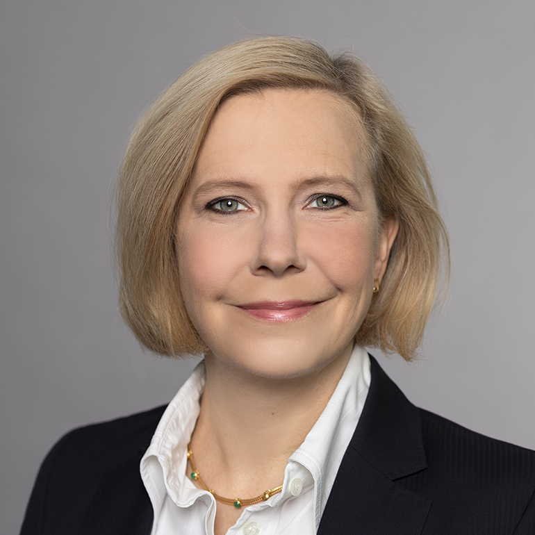 Portrait einer Frau mit blonden, schulterlangen Haaren. Sie trägt ein schwarzes Jackett und eine weiße Bluse. Ihr Gesicht strahlt Freundlichkeit aus, mit einem leichten Lächeln. Der Hintergrund ist grau.