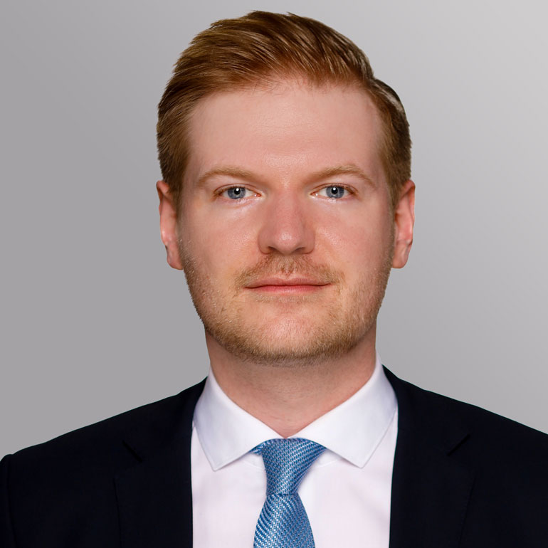 Porträt eines Mannes mit blonden Haaren, einem kurzen Bart und blauen Augen. Er trägt einen dunklen Anzug mit einem weißen Hemd und einer blauen Krawatte. Der Hintergrund ist grau.