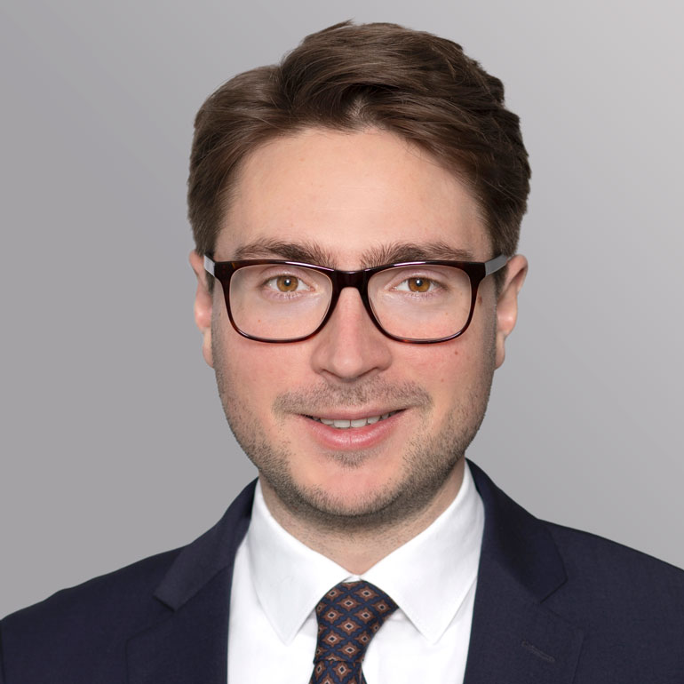 Porträt eines Mannes mit kurz geschnittenen, braunen Haaren und einer Brille. Er trägt einen dunklen Anzug mit einer hellen Krawatte und lächelt freundlich. Der Hintergrund ist grau.