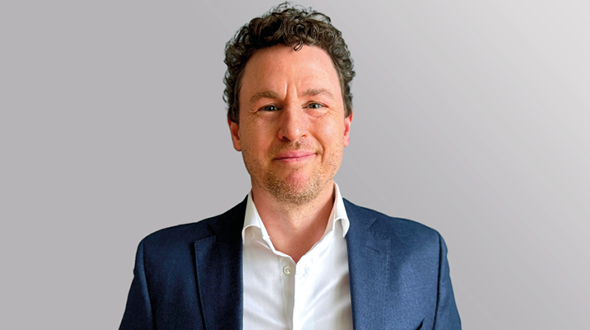 Ein lächelnder Mann mit lockigem, braunem Haar trägt ein dunkelblaues Sakko und ein weißes Hemd. Er steht vor einem hellen, einfarbigen Hintergrund und blickt direkt in die Kamera.