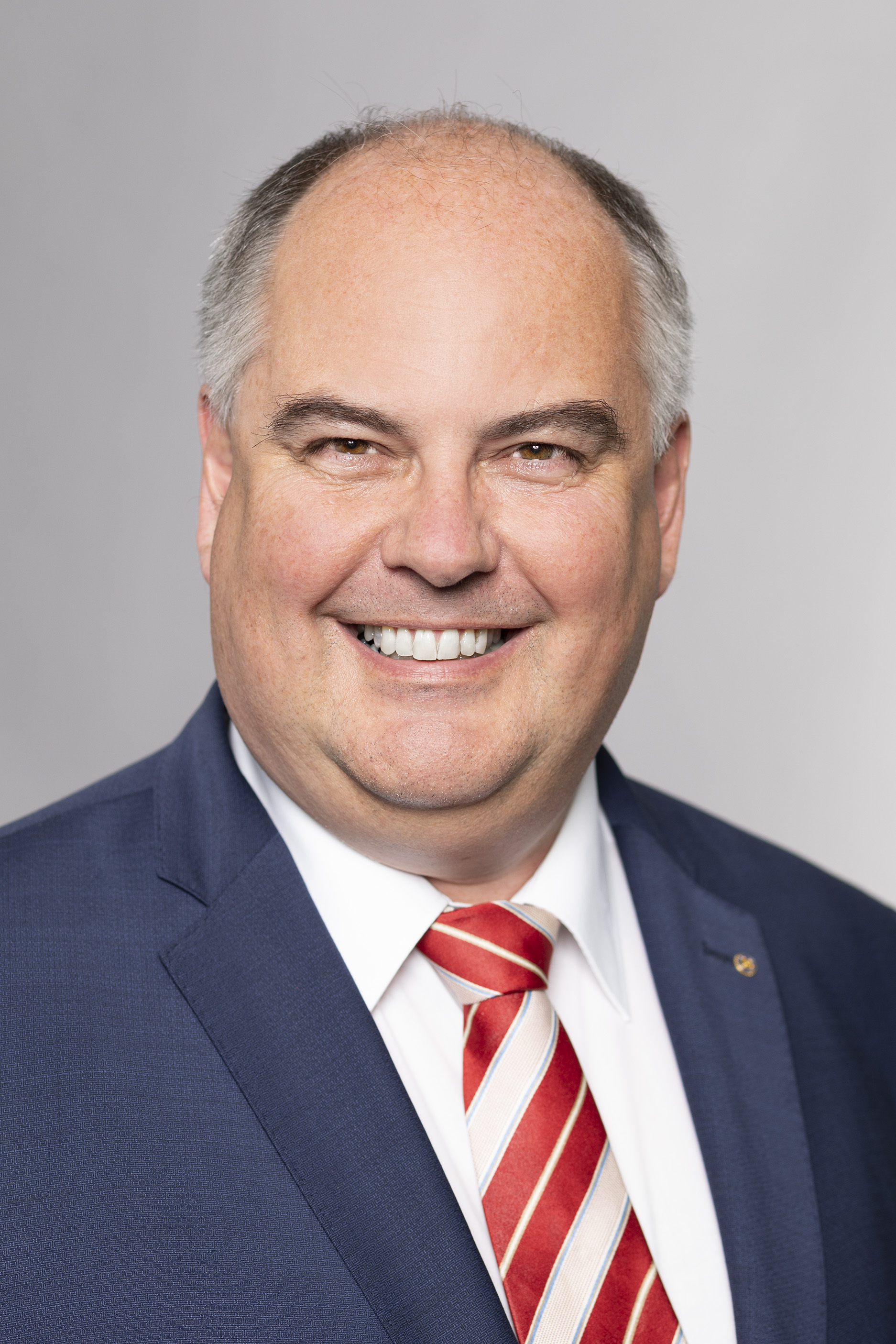 A middle-aged man smiles warmly, wearing a dark suit with a white shirt and a red-striped tie. His hair is gray with a balding pattern. The background is light gray, emphasizing his friendly expression.