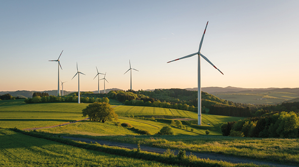 Windkraftanlagen auf einer grünen Wiese am Abend. Im Hintergrund sind sanfte Hügel und Wälder sichtbar, während die Sonne untergeht und eine warme Atmosphäre schafft.