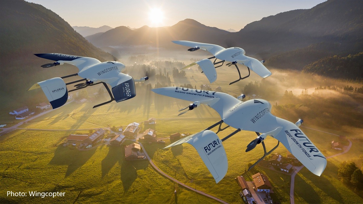 Three drones are flying over a picturesque mountainous landscape, surrounded by mist and lush greenery. Below, small farms and houses are visible against the backdrop of rolling hills and trees. The scene captures a blend of technology and nature during early morning light.