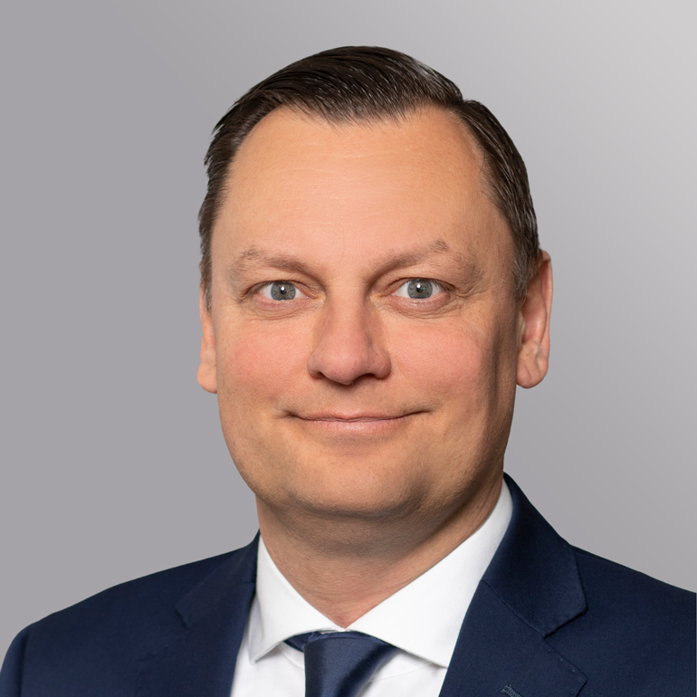 Porträt eines Mannes mit kurzen, dunkelblonden Haaren, Hauttyp hell. Er trägt einen dunklen Anzug und eine helle Krawatte. Der Mann lächelt leicht in die Kamera, vor einem neutralen, grauen Hintergrund. Sein Ausdruck wirkt freundlich und professionell.