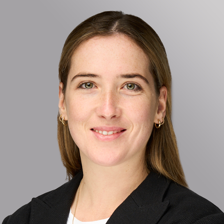 Ein Portrait einer Frau mit langen, glatten Haaren und einem freundlichen Lächeln. Sie trägt einen schwarzen Blazer und hat goldene Ohrringe. Der Hintergrund ist in sanften Grautönen gehalten.