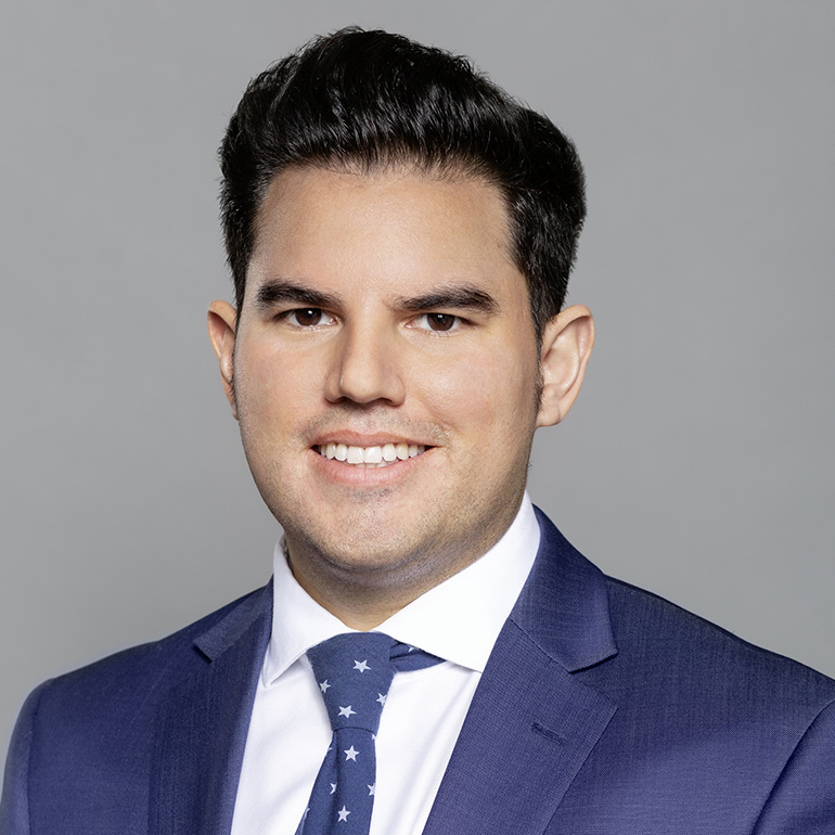 Ein lächelnder junger Mann mit dunklen Haaren trägt einen dunkelblauen Anzug und eine hellblaue Krawatte mit Sternen. Der Hintergrund ist grau. Sein Gesicht ist gut beleuchtet, und er schaut direkt in die Kamera.