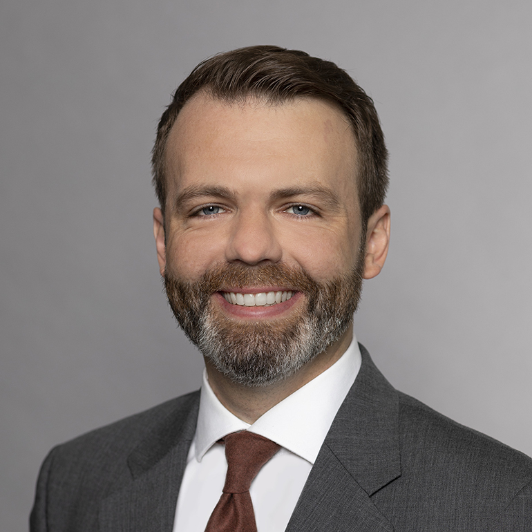 Das Bild zeigt einen lächelnden Mann mit Bart, der einen grauen Anzug und eine weiße Hemdbluse trägt, sowie eine braune Krawatte. Der Hintergrund ist grau. Die Person wirkt freundlich und professionell.