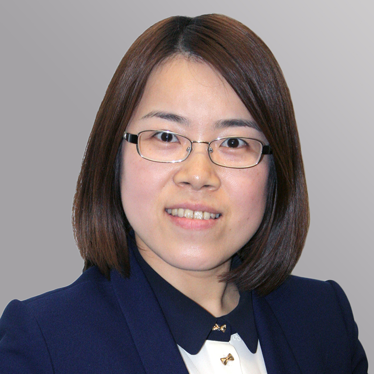 Porträt einer Frau mit schulterlangen, glatten Haaren, die eine Brille trägt. Sie lächelt freundlich und hat ein blaues Sakko sowie ein helles Oberteil an. Der Hintergrund ist grau.