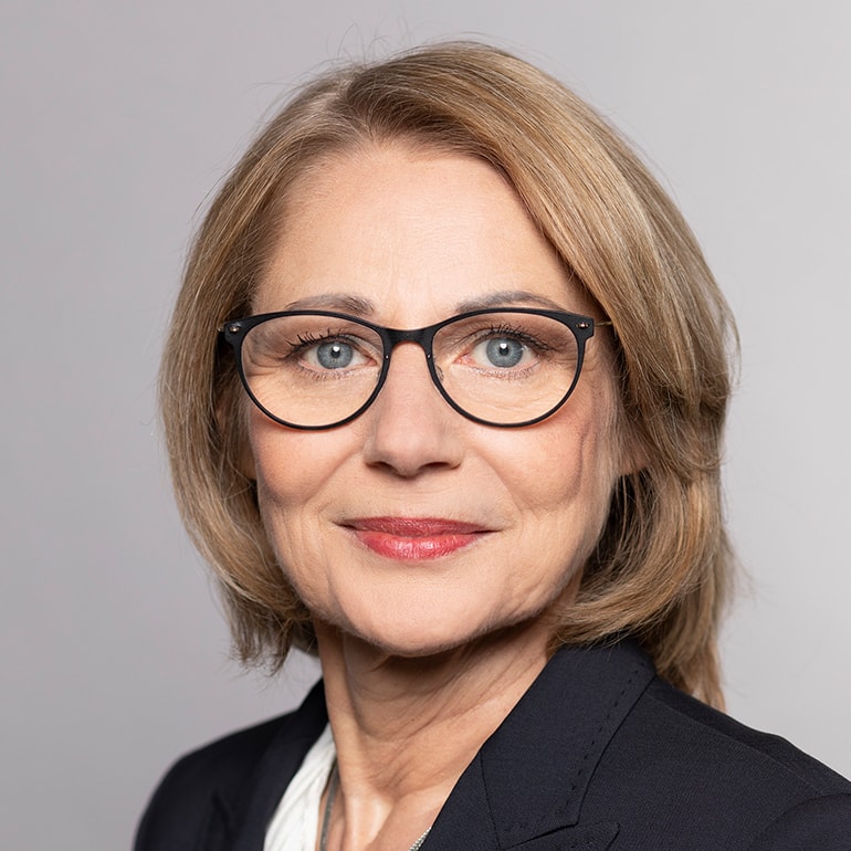 Porträt einer Frau mittleren Alters mit blonden, schulterlangen Haaren und einer Brille. Sie trägt einen dunklen Blazer und hat einen freundlichen, selbstbewussten Gesichtsausdruck. Der Hintergrund ist neutral und hell.