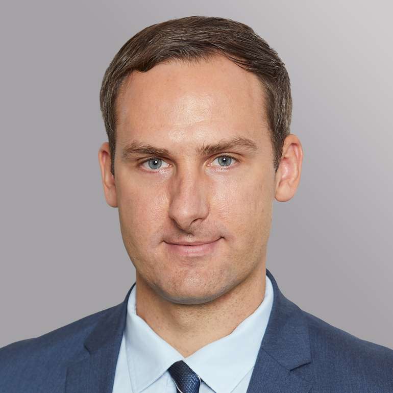 Porträt eines Mannes mit kurzen, braunen Haaren und blauen Augen. Er trägt einen dunklen Anzug und ein helles Hemd sowie eine Krawatte. Der Hintergrund ist einfarbig grau, was den Fokus auf den Mann legt. Sein Ausdruck ist neutral und professionell.