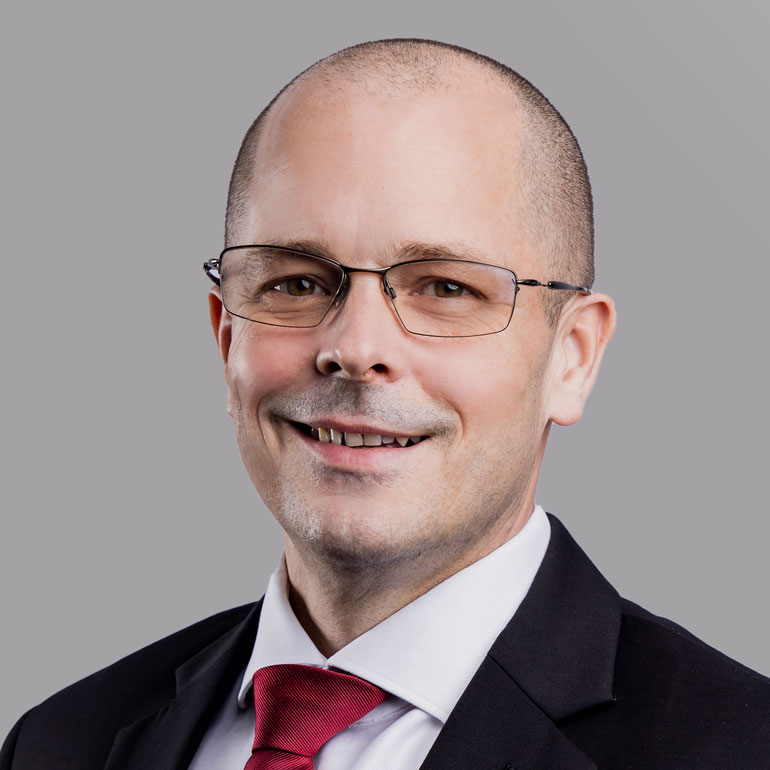 Porträt eines Mannes mit Glatze und Brille, der freundlich lächelt. Er trägt einen schwarzen Anzug mit einem weißen Hemd und einer roten Krawatte. Der Hintergrund ist grau und neutral gehalten.