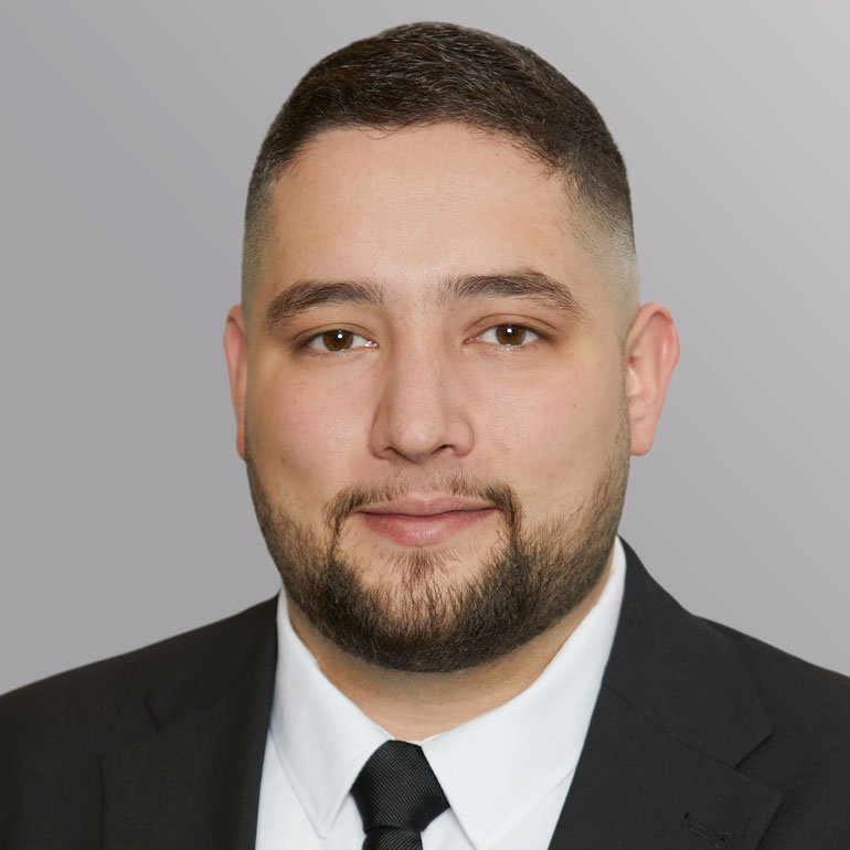 Porträt eines Mannes mit kurzem, dunklem Haar und Bart. Er trägt einen schwarzen Anzug und eine weiße Hemdbluse mit Krawatte. Der Hintergrund ist hellgrau. Der Mann lächelt und schaut direkt in die Kamera.