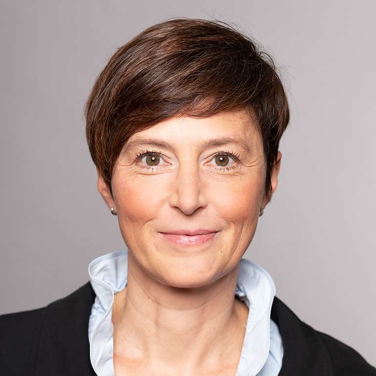 Porträt einer Frau mit kurzem, braunem Haar und einem freundlichen Lächeln. Sie trägt ein schwarzes Oberteil mit einem hellblauen Kragen. Der Hintergrund ist einfarbig und hell.