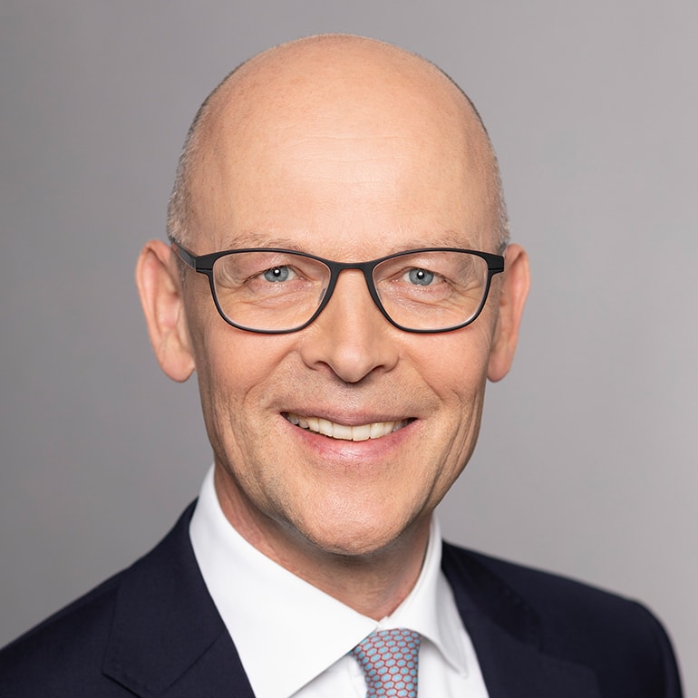 Porträt eines Mannes mit Glatze, der eine Brille trägt. Er lächelt freundlich und ist in einem formellen Outfit gekleidet, einschließlich eines Anzugs und einer Krawatte. Der Hintergrund ist neutral und hellgrau.