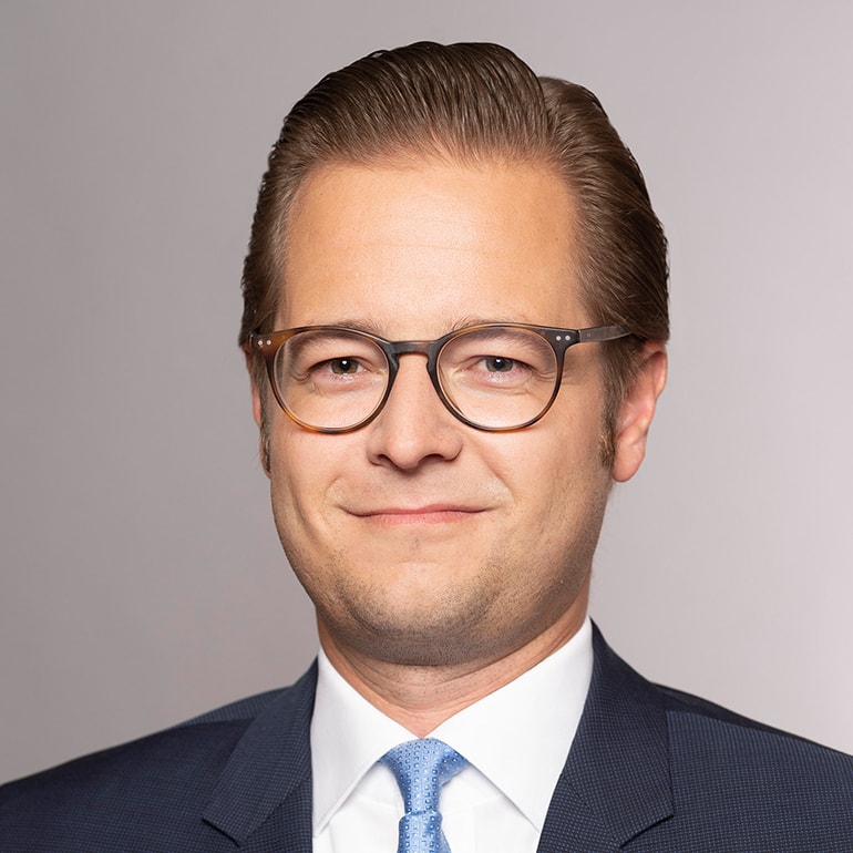 Ein männlicher Geschäftsmann mit Brille lächelt in die Kamera. Er trägt einen dunklen Anzug und eine hellblaue Krawatte. Der Hintergrund ist einheitlich grau, wodurch sein Gesicht und die Kleidung gut zur Geltung kommen.