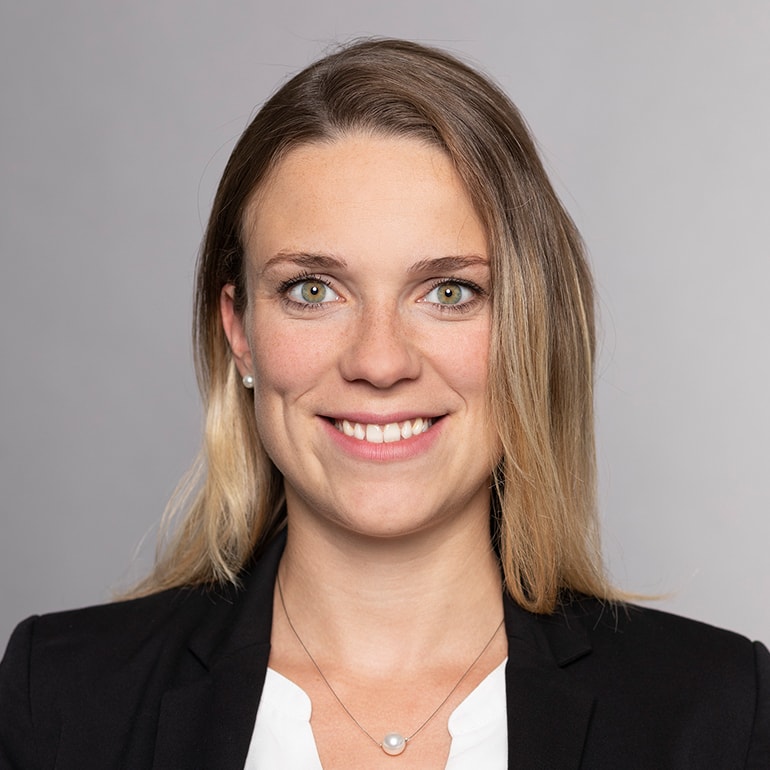 Ein Porträt einer Frau mit langen, blonden Haaren und einem freundlichen Lächeln. Sie trägt einen schwarzen Blazer und eine weiße Bluse. Der Hintergrund ist neutral und sorgt dafür, dass der Fokus auf ihrem Gesicht liegt.