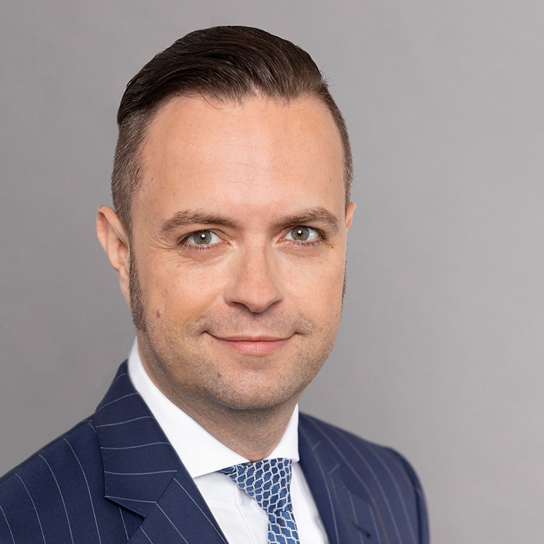 Ein Mann mit kurzem, glattem Haar und einem leichten Bart lächelt in die Kamera. Er trägt einen dunkelblauen Anzug mit feinen Streifen und eine helle Krawatte mit Muster. Der Hintergrund ist grau.