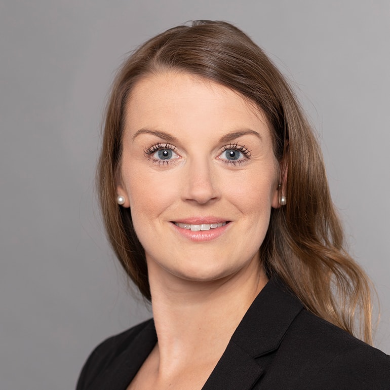 Ein Porträt einer Frau mit langen, braunen Haaren und blauen Augen. Sie trägt einen schwarzen Blazer und hat Perlenohrringe. Sie lächelt freundlich vor einem grauen Hintergrund.