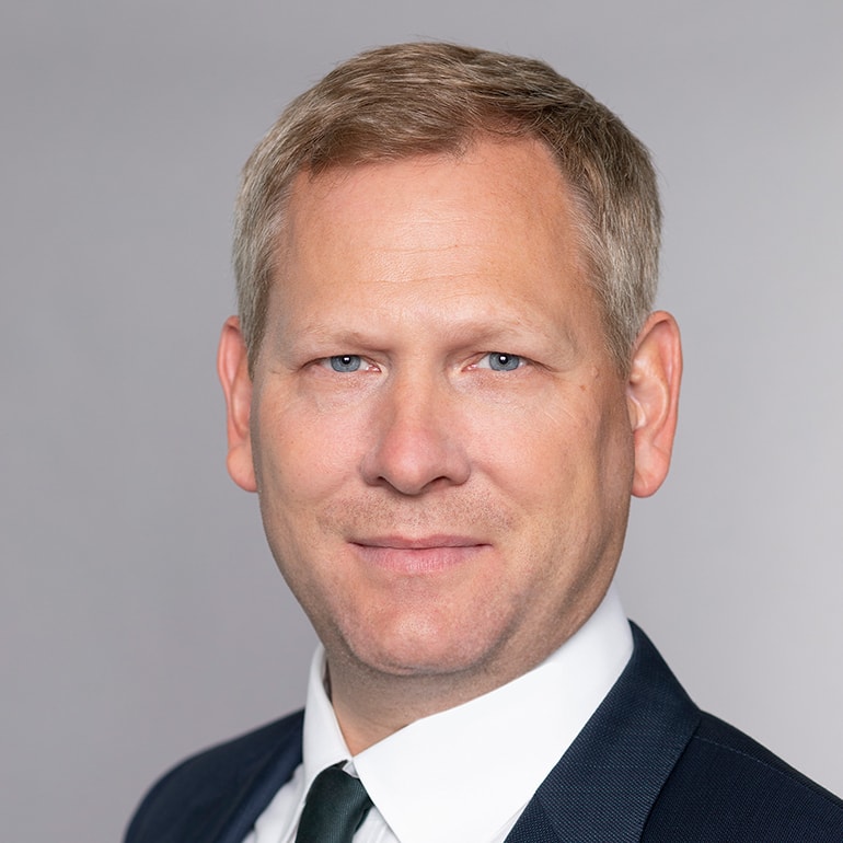 Porträt eines Mannes mit kurzen blonden Haaren und blauen Augen. Er trägt einen dunklen Anzug mit Krawatte und hat einen neutralen Ausdruck auf dem Gesicht. Der Hintergrund ist in einem einfarbigen Grau gehalten.