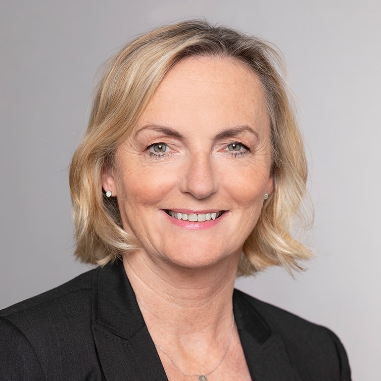 Porträt einer lächelnden Frau mit blonden, schulterlangen Haaren, die einen schwarzen Blazer trägt. Der Hintergrund ist neutral und hell. Die Frau sieht freundlich und selbstbewusst aus.